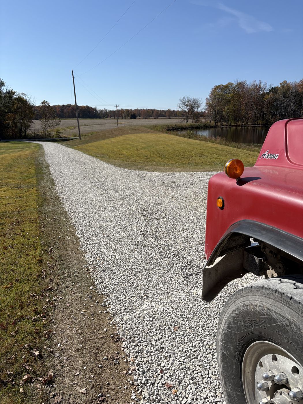 A freshly finished white gravel driveway curving toward a pond, with the front of Aaron's red dump truck at the edge of the frame